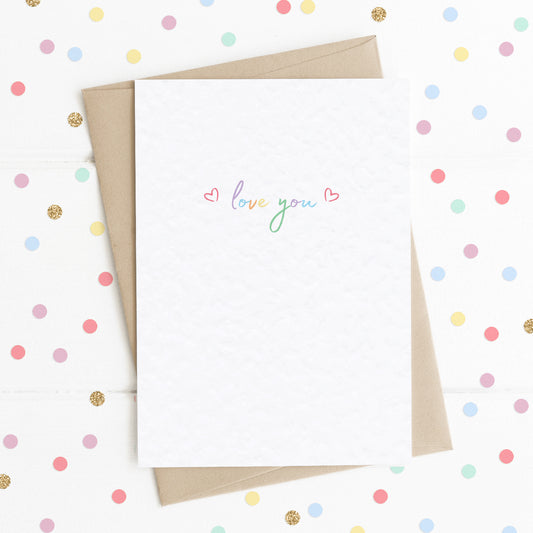 A cute A6 love card with the message "Love You" in colourful rainbow type, with two reds hearts either side.